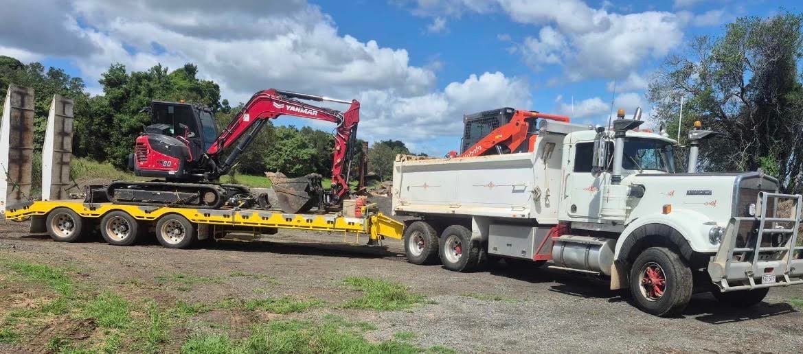 A red excavator and orange skid steer loader transported on yellow and black trailers hitched to a white truck at a construction site